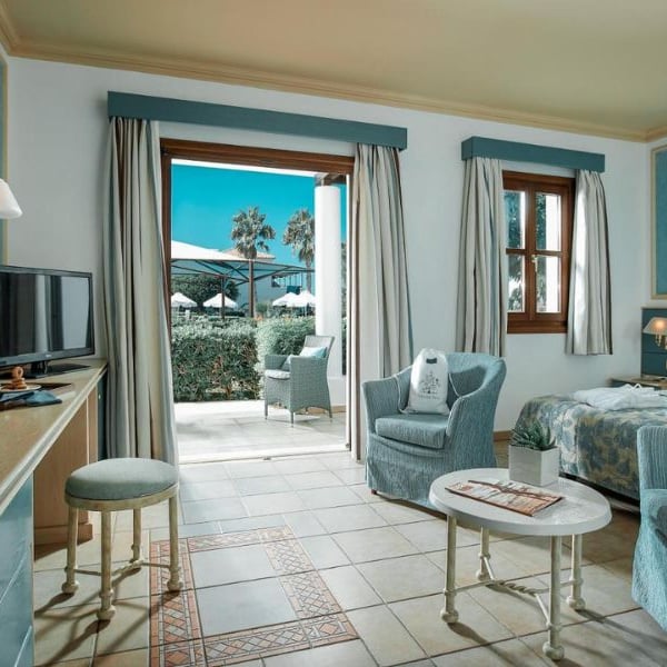 Luxurious hotel room with blue and white decor at Royal Mare, featuring a twin bed, armchair, TV, and garden view with palm trees and umbrellas.