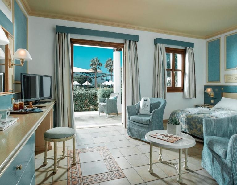 Luxurious hotel room with blue and white decor at Royal Mare, featuring a twin bed, armchair, TV, and garden view with palm trees and umbrellas.