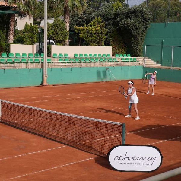 Two women playing tennis on a clay court at Royal Mare, with one serving and the other at the baseline. Surroundings include green fences and seating.