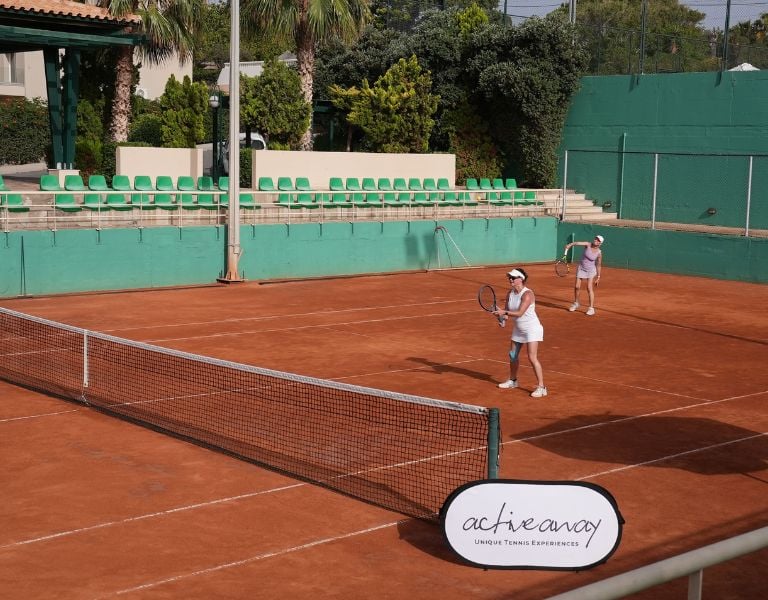 Two women playing tennis on a clay court at Royal Mare, with one serving and the other at the baseline. Surroundings include green fences and seating.
