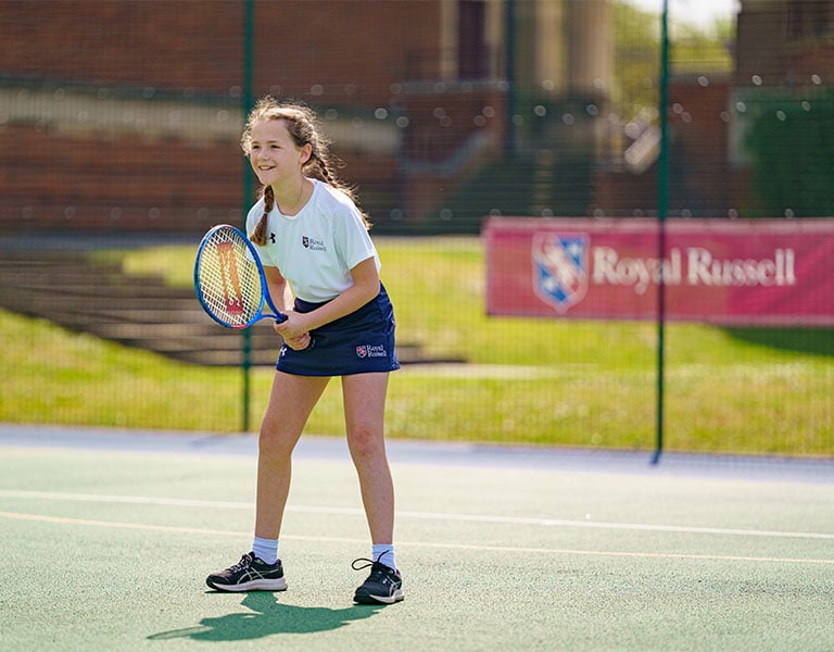 Royal Russell School student in ready position on outdoor tennis court with racket, wearing sports gear with school branding in the background.