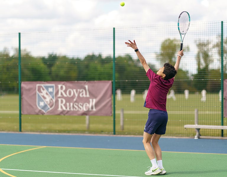 Student serving tennis on the Royal Russell School tennis court with school branding and a scenic backdrop.