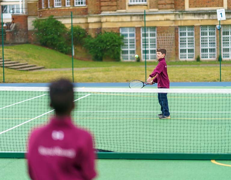 Two students playing tennis on an outdoor court at Royal Russell School, with a brick building in the background.