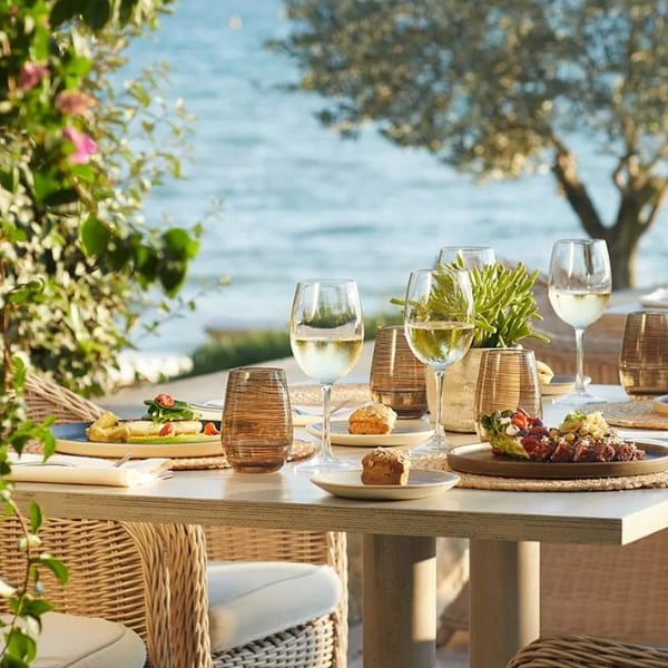 Outdoor dining table at Sani Beach with gourmet dishes, glasses of white wine, wicker chairs, and ocean view in the background.