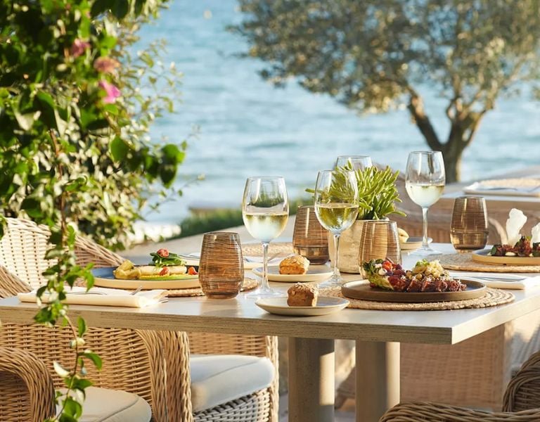 Outdoor dining table at Sani Beach with gourmet dishes, glasses of white wine, wicker chairs, and ocean view in the background.