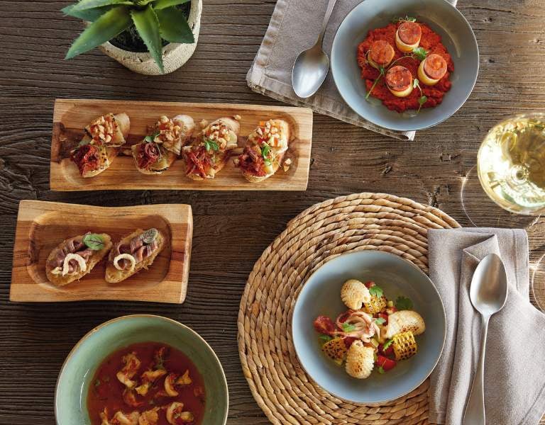 Mediterranean meal at Sani Beach with bruschetta, tomato-based dish, soup, and grilled shellfish on a rustic wooden table setting with a glass of white wine.