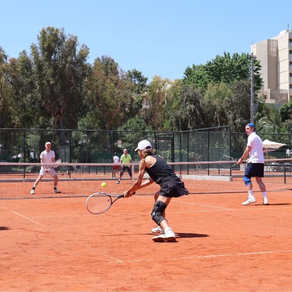 Tennis players on a red clay court at Sani Beach Resort with trees and buildings in the background.