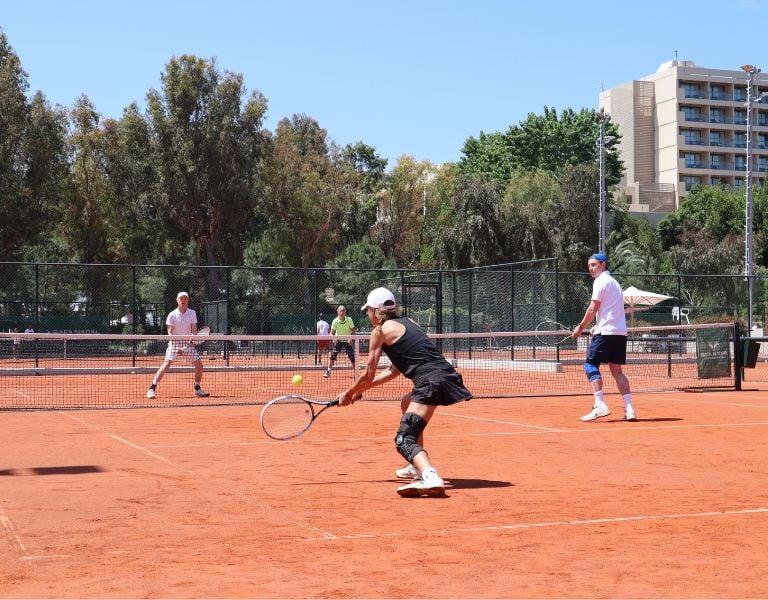 Tennis players on a red clay court at Sani Beach Resort with trees and buildings in the background.