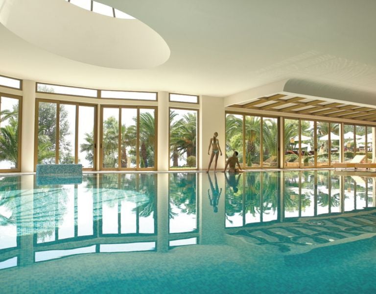 Luxurious indoor pool at Sani Beach with large windows showcasing views of palm trees and reflective blue water.