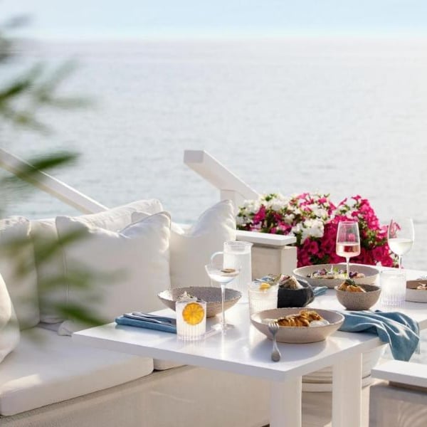 Outdoor dining area at Sani Beach with a sea view, white couch, and a table beautifully set with glassware and dishes, surrounded by vibrant pink flowers.