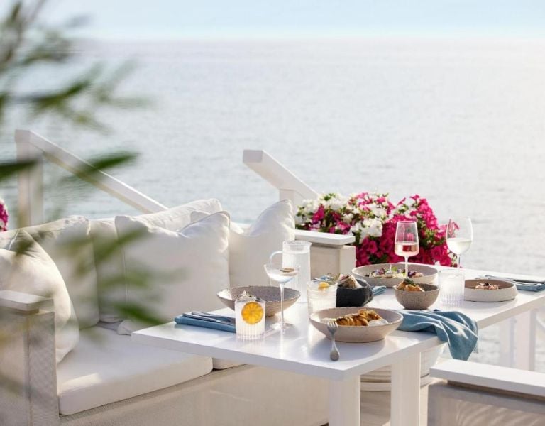 Outdoor dining area at Sani Beach with a sea view, white couch, and a table beautifully set with glassware and dishes, surrounded by vibrant pink flowers.