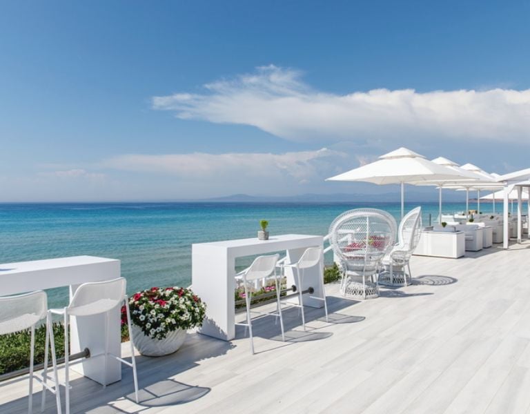 Outdoor seating at Sani Beach Resort, with white tables and umbrellas overlooking the turquoise sea.