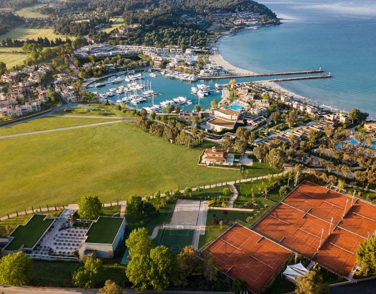 Aerial view of Sani Resort, Halkidiki, featuring a marina, sandy beach, tennis courts, and lush landscapes by the Aegean Sea.