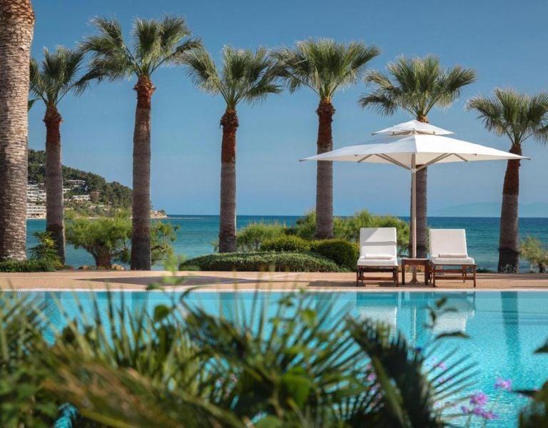 Sani Beach pool lounge with two sun loungers, umbrella, palm trees, a pool, and the sea in the background.