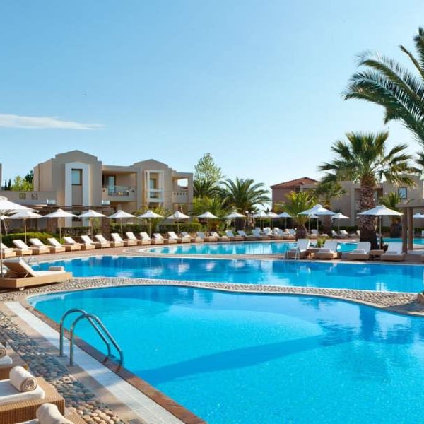 Luxurious outdoor pool at Sani Beach with sun loungers, palm trees, and modern buildings in the background.