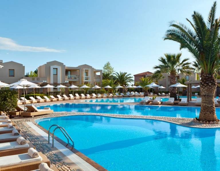 Luxurious outdoor pool at Sani Beach with sun loungers, palm trees, and modern buildings in the background.