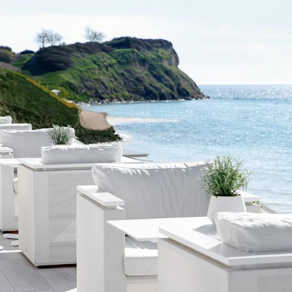 Outdoor dining area at Sani Beach with white seating and tables, overlooking the sea and a grassy hill under a clear sky.
