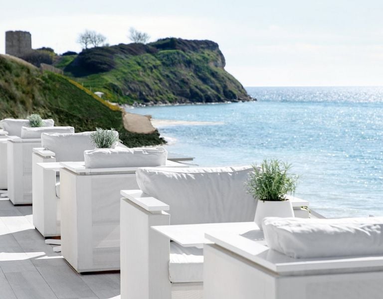 Outdoor dining area at Sani Beach with white seating and tables, overlooking the sea and a grassy hill under a clear sky.