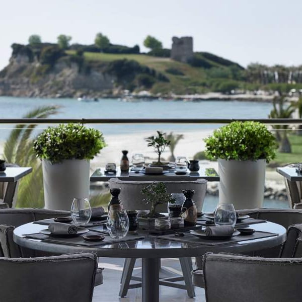 Outdoor terrace of Sani Beach Resort restaurant with elegantly set tables, overlooking a sandy beach, blue water, and an island with greenery and a historic tower.