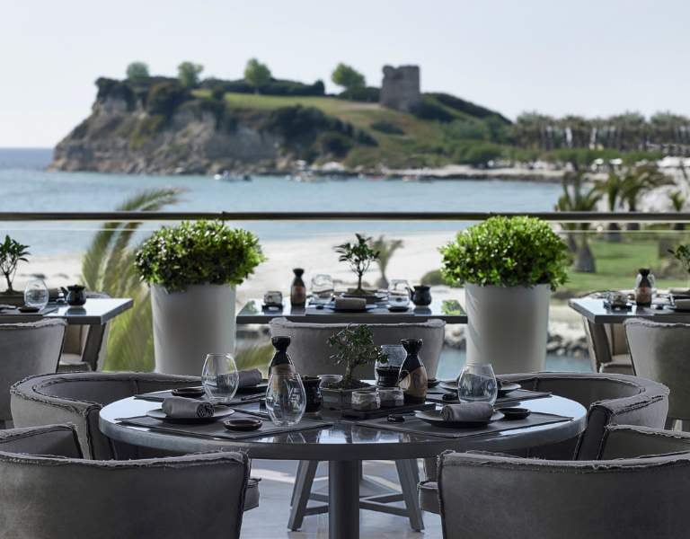 Outdoor terrace of Sani Beach Resort restaurant with elegantly set tables, overlooking a sandy beach, blue water, and an island with greenery and a historic tower.