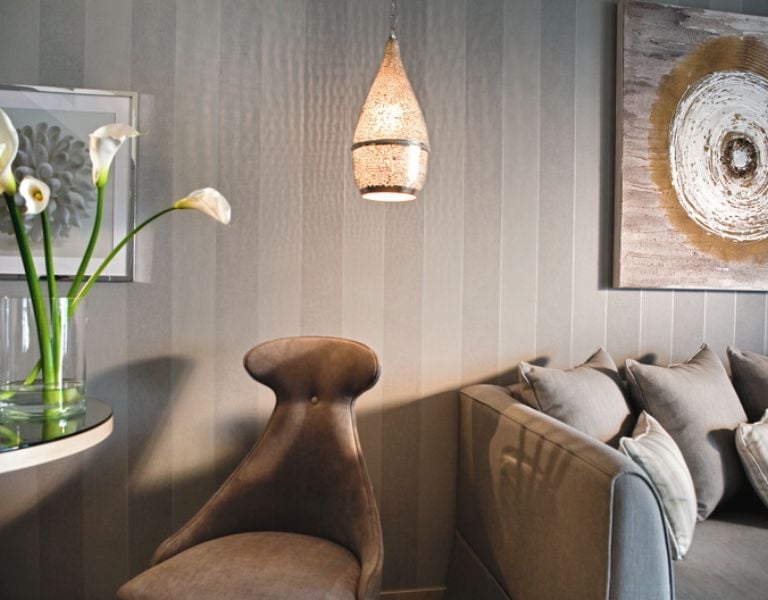 Interior view of a Sani Beach room with grey striped wall, plush grey chair, table with calla lilies, sofa with cushions, and decorative art with hanging light.