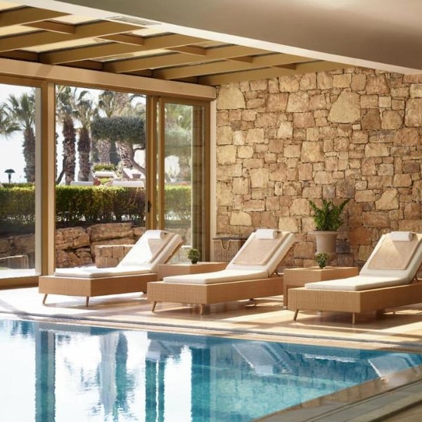 Spa lounge at Sani Beach with sun loungers by an indoor pool, surrounded by large glass doors showing garden and palms.