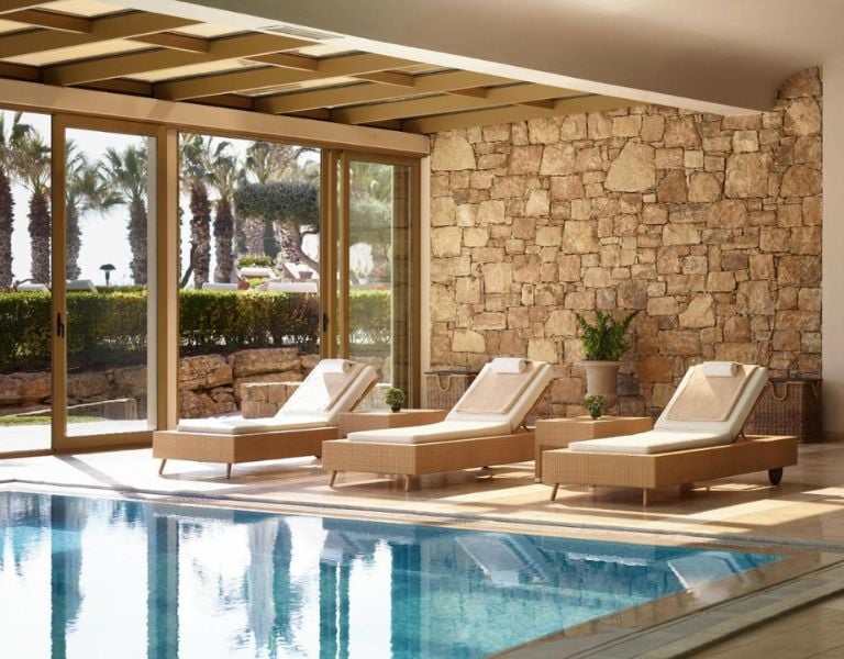 Spa lounge at Sani Beach with sun loungers by an indoor pool, surrounded by large glass doors showing garden and palms.
