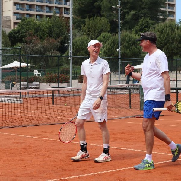 Two men in athletic gear playing tennis on a clay court at Sani Beach resort, surrounded by greenery and resort buildings.