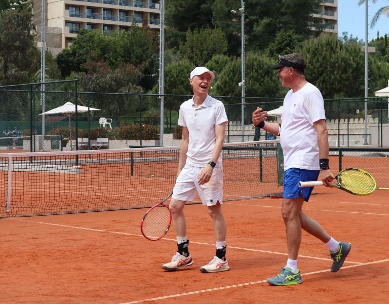 Two men in athletic gear playing tennis on a clay court at Sani Beach resort, surrounded by greenery and resort buildings.