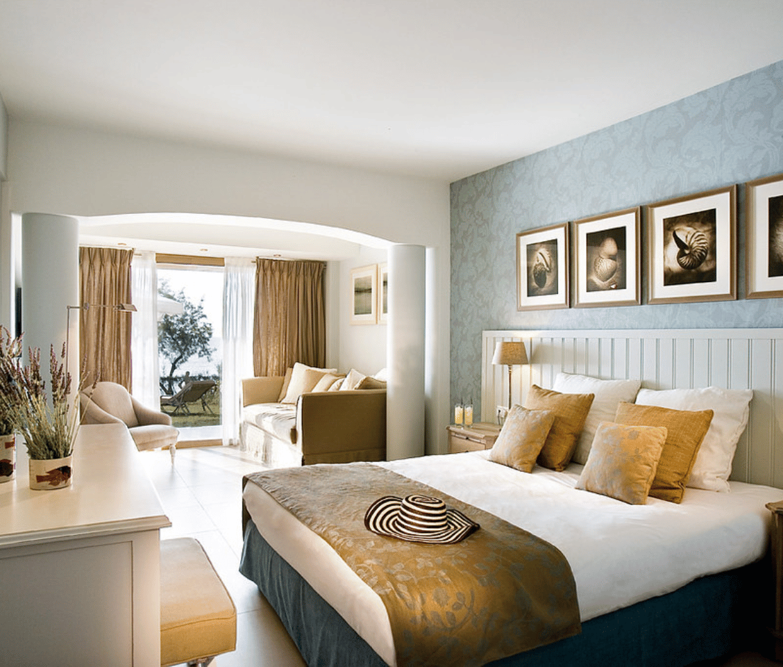 Interior of Sani Beach Junior Suite with a double bed, decorative cushions, seating area, and view of the garden through large windows.