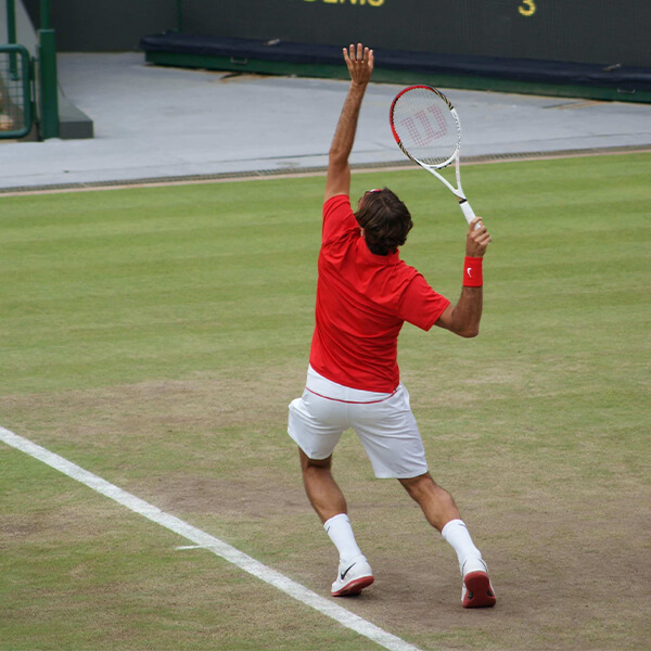 Tennis player in red shirt and white shorts serving on a grass tennis court, racquet raised overhead.