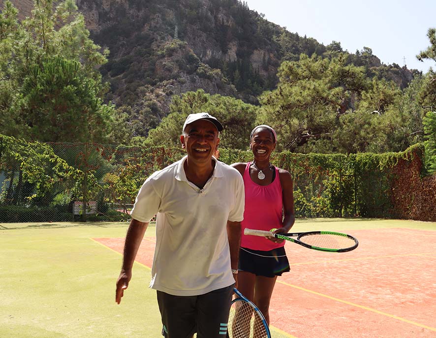 Man and woman smiling on a green tennis court with rackets, surrounded by trees and mountains, enjoying a tennis holiday.