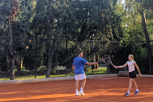 Two tennis players, a man and a woman, shaking hands over the net on a clay court amidst trees.