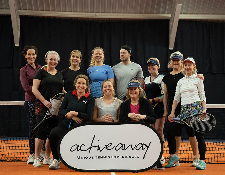 Group of smiling tennis players on an indoor court, holding rackets, with an 'active away' sign for a tennis clinic.