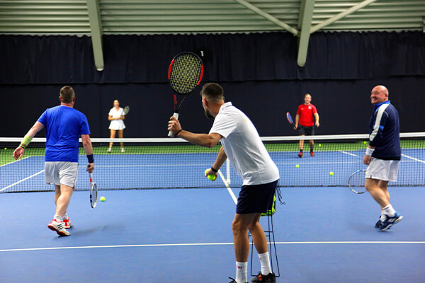 Group tennis clinic in an indoor setting with four adults playing doubles, focusing on improving their game on a blue court.
