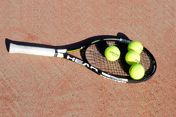 Tennis racket with four tennis balls on a clay court, featuring a Head brand racket.