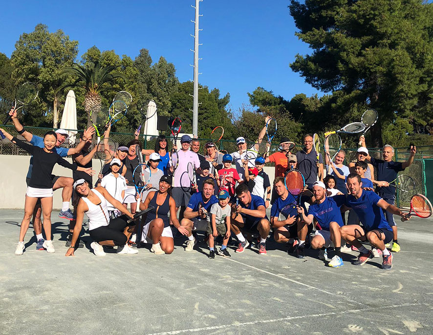 Group of adults and children posing on a tennis court, holding tennis rackets, under a blue sky with trees in the background.