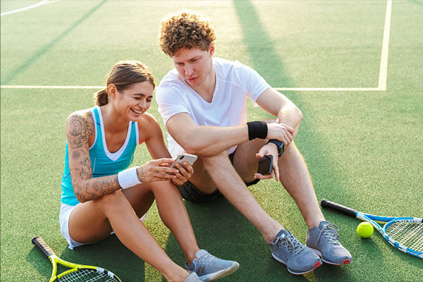 Two tennis players sitting on a court, smiling and looking at a smartphone, with tennis rackets beside them.