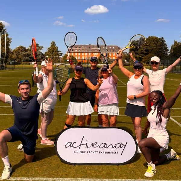 Active Away tennis team on grass courts at The Lensbury, posing with rackets raised, with the historic building in the background.