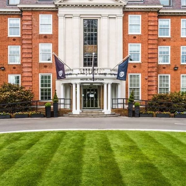 Front view of The Lensbury hotel entrance, showcasing its red-brick facade, orderly lawns, and flags above the revolving door entrance.
