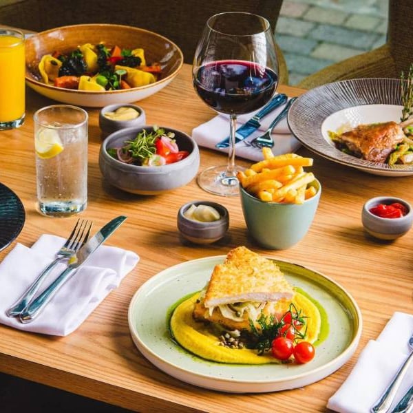 Dining table at The Lensbury featuring gourmet dishes such as roasted chicken, pasta, salad, and wine.