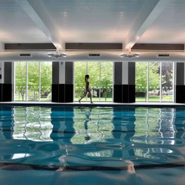 Indoor pool at The Lensbury with a person walking alongside and large windows displaying green trees outside.