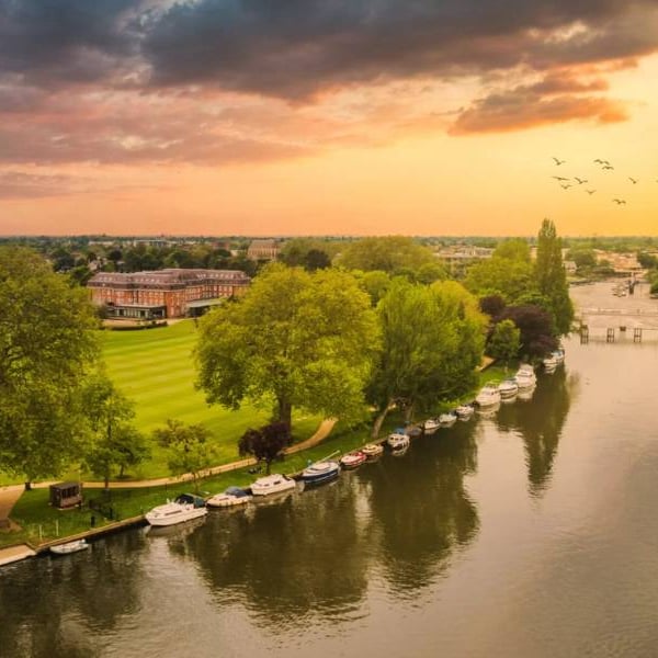 Aerial view of The Lensbury resort on the River Thames, with boats, lush greenery, and a sunset sky.