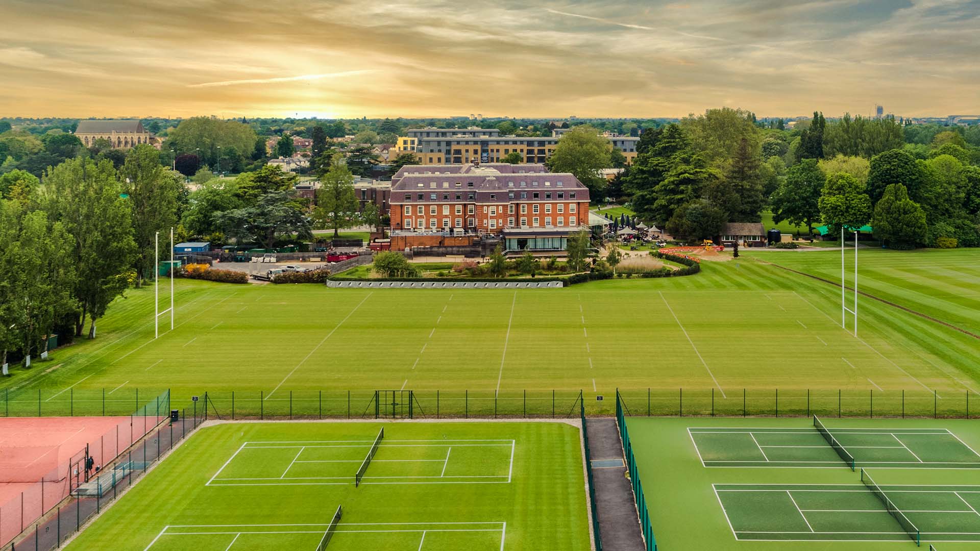Aerial view of The Lensbury sports facilities with tennis courts, rugby fields, and a clubhouse set against a sunset backdrop.
