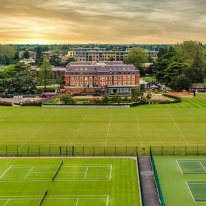 Aerial view of The Lensbury sports facilities with tennis courts, rugby fields, and a clubhouse set against a sunset backdrop.