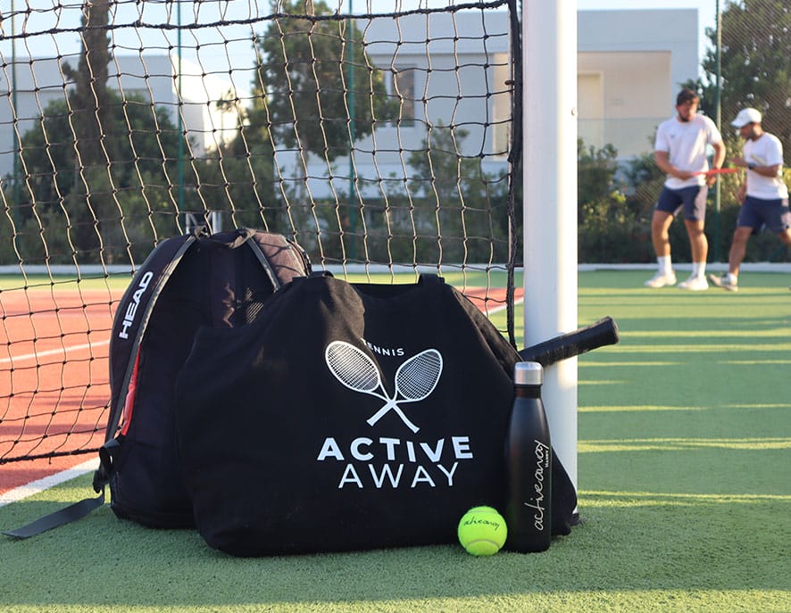 Active Away tennis bag with racquet, ball, and water bottle on a court, with two players practising in the background.