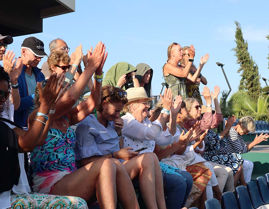Crowd in outdoor tennis stadium clapping and cheering during a match, with a mix of men and women in summer attire, blue sky and greenery in the background.