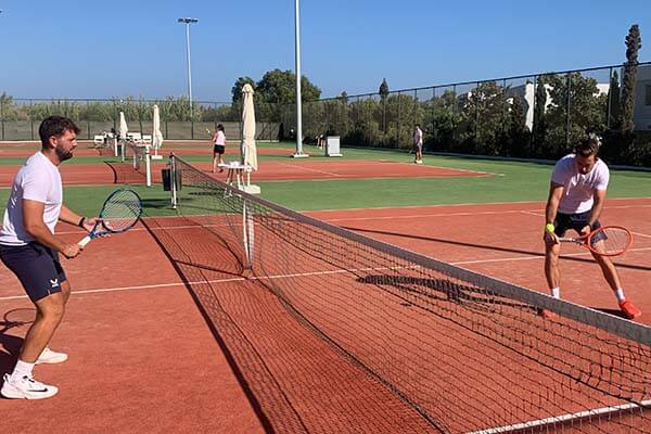 Two male tennis players warming up on a clay court, focusing on net play under a clear blue sky.