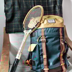 Blue and tan rucksack with leather straps and a vintage wooden badminton racket on a wooden chair with a plaid blanket draped over it.