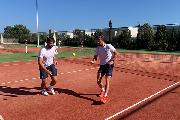 Two tennis players practising volleys on a clay court, with a tennis ball in mid-air between them, under a clear blue sky.
